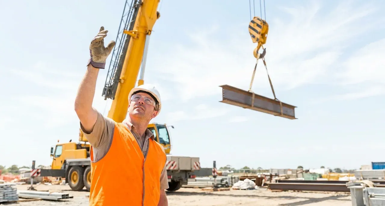 Rigger in hi-vis directing a suspended load with hand signals below a mobile crane