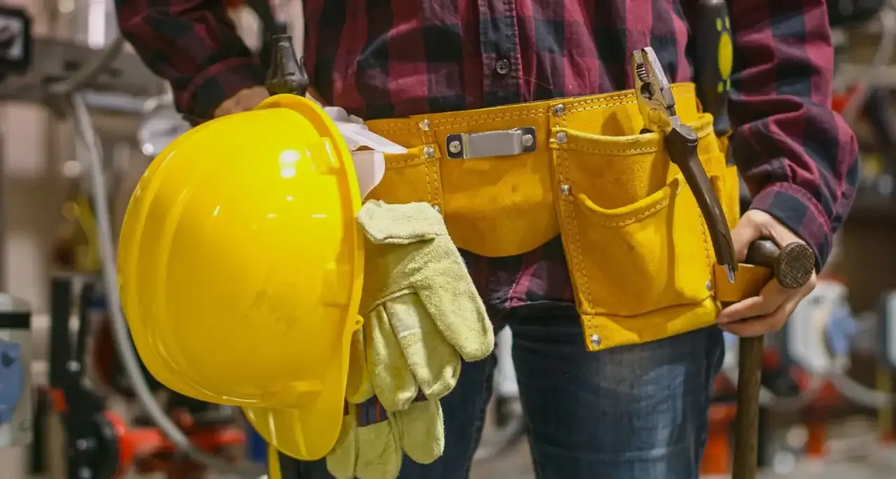 Worker holding a hard hat and gloves with a tool belt fitted